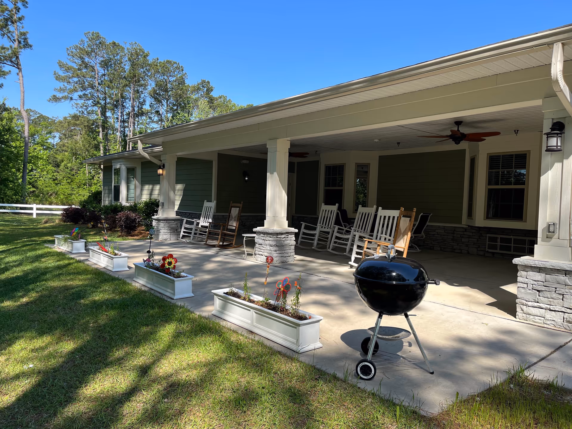 Covered front porch of an assisted living facility with rocking chairs, a charcoal grill, and flower planters on a lawn.