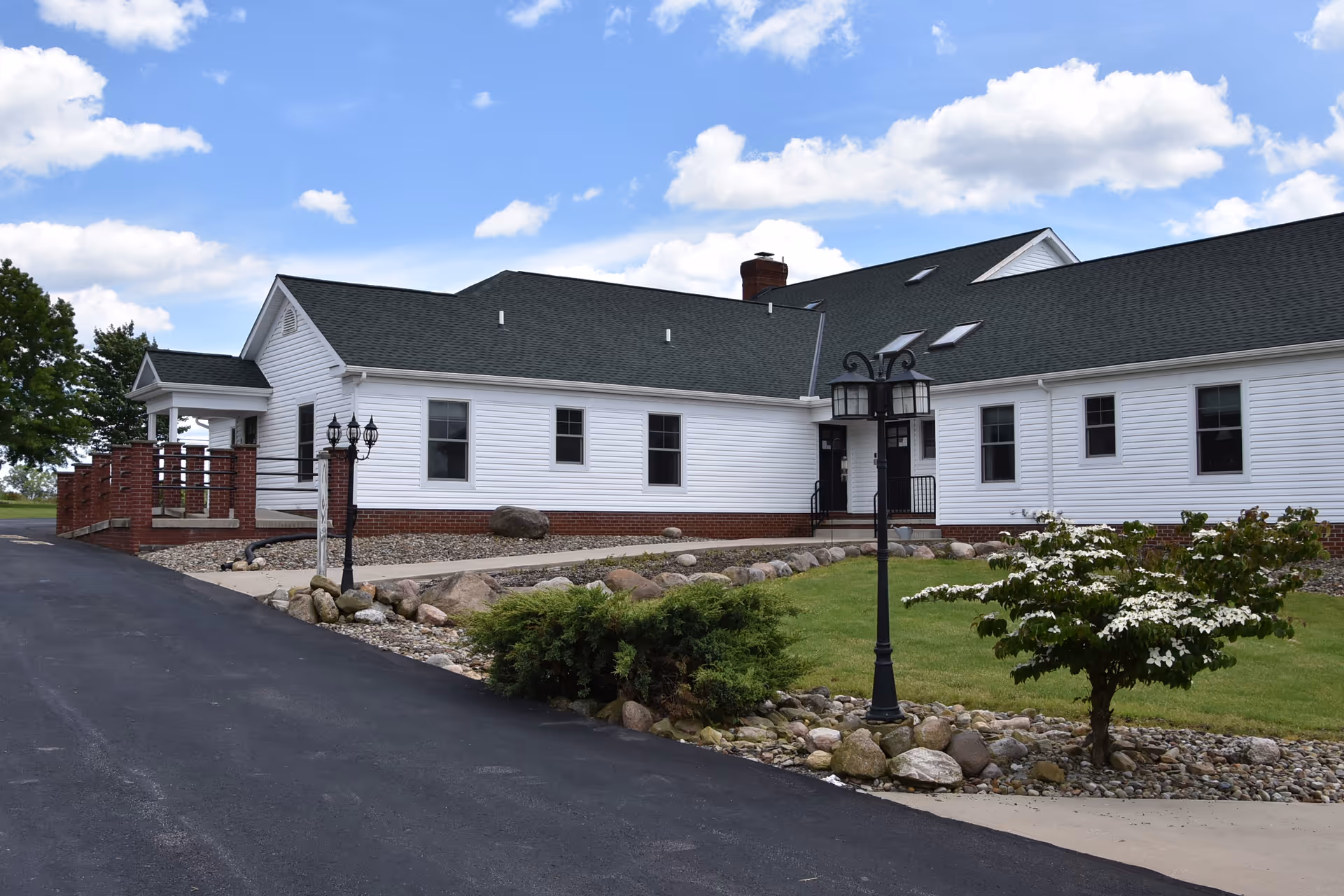 Exterior view of a single-story white building with a dark roof, surrounded by a paved driveway, green lawn, rocks, and small trees under a partly cloudy blue sky.