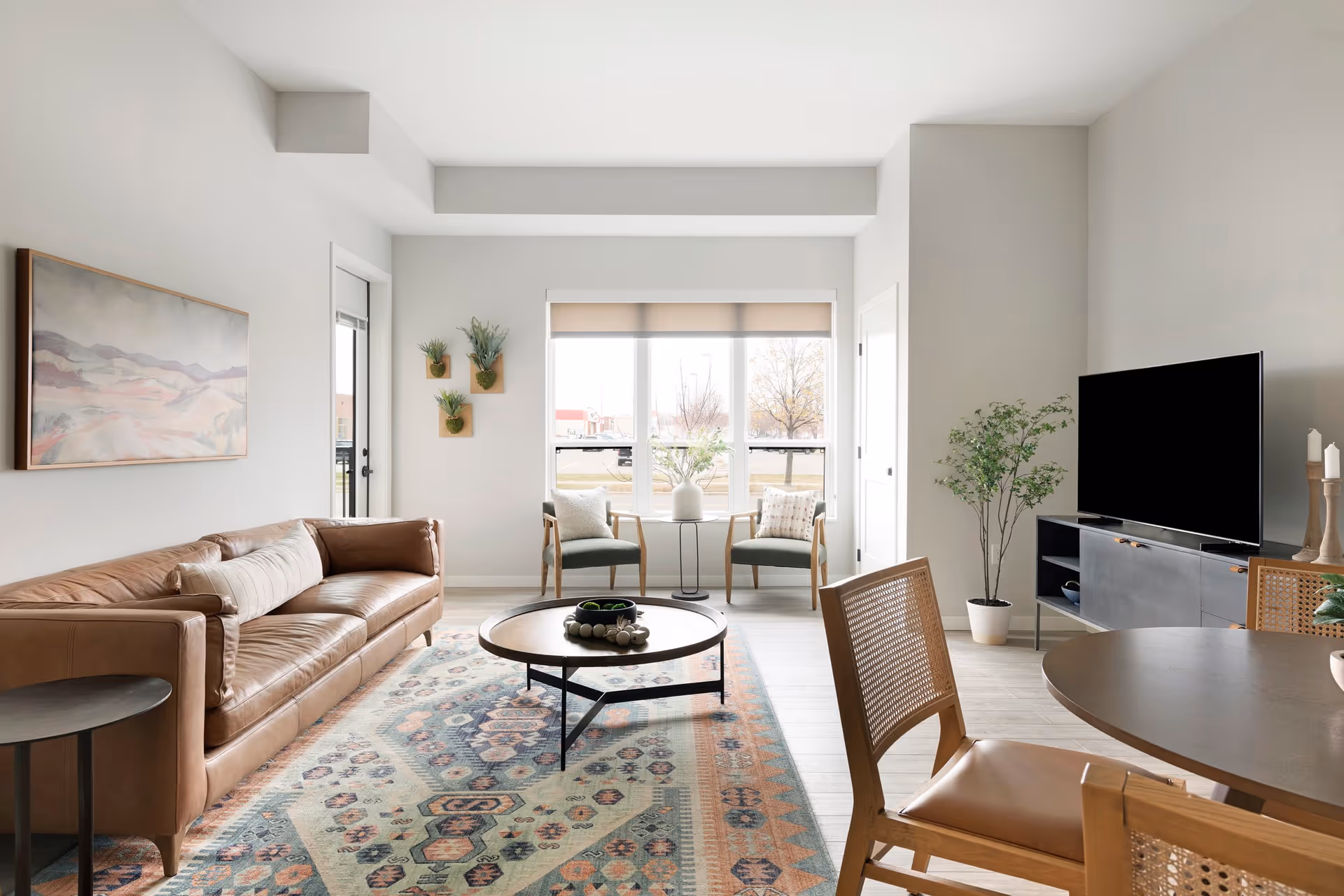 A modern living room with a brown leather sofa, a round wooden coffee table on a patterned rug, two green armchairs by a large window, a TV on a black media console, and a round dining table with wooden chairs in the foreground. The walls are light gray with minimal decor including a landscape painting and small wall-mounted plants.