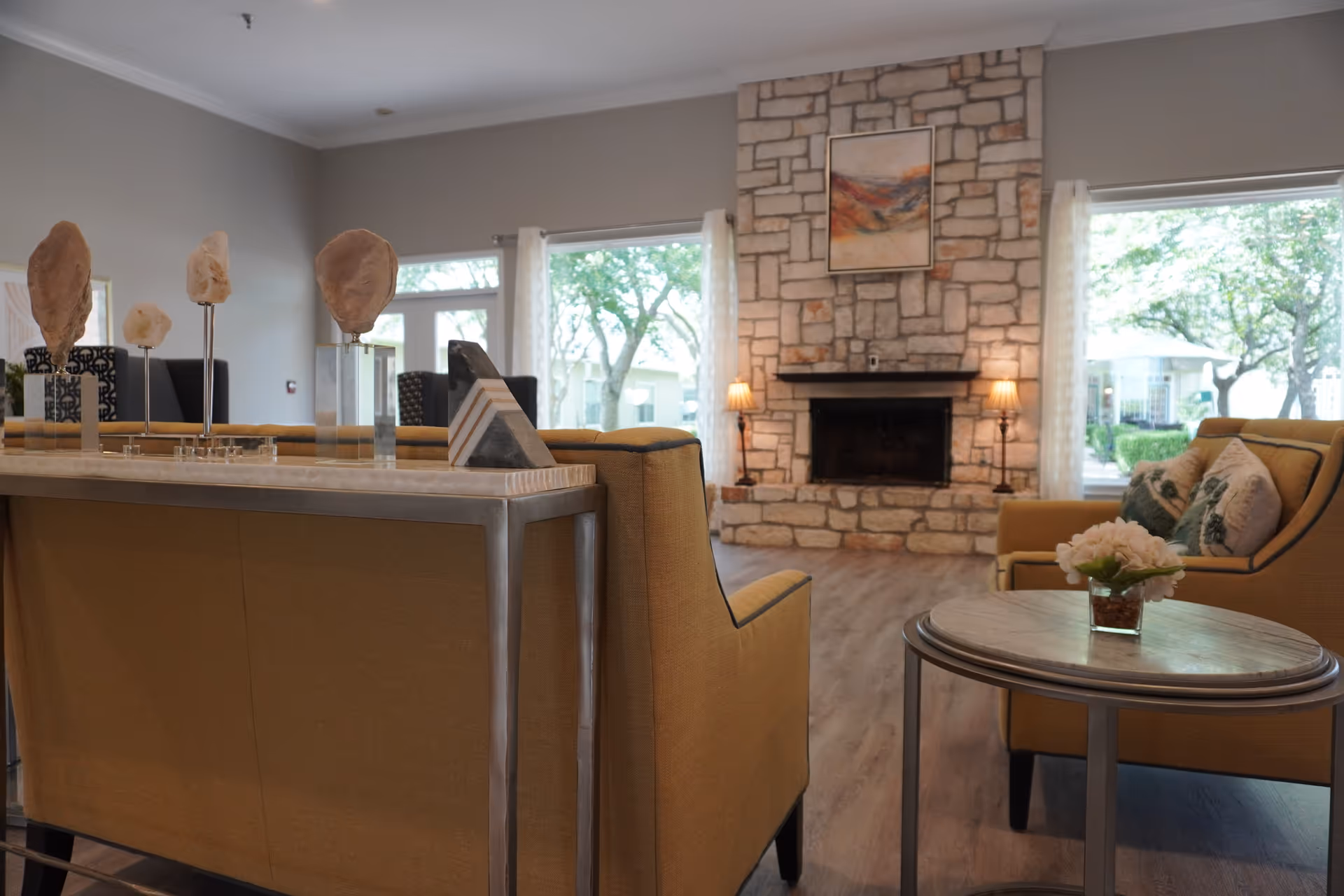 A cozy living room with mustard yellow armchairs and a matching sofa. A stone fireplace with a painting above it is centered on the far wall, flanked by two lamps. Large windows with sheer white curtains let in natural light and show green trees outside. A round wooden side table with a small flower arrangement is in the foreground.