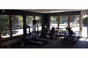 A rehabilitation room with exercise equipment including treadmills and therapy tables, surrounded by large windows letting in natural light and providing a view of trees outside.