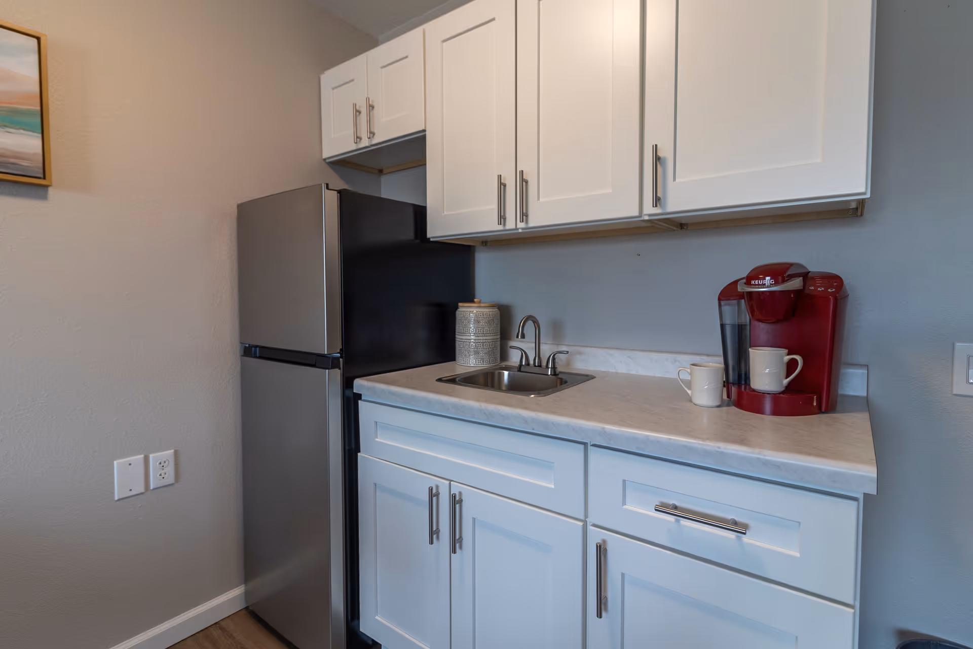 A small kitchen area with white cabinets, a stainless steel refrigerator, a sink with a faucet, a red Keurig coffee maker, two white mugs, and a decorative jar on the countertop. A framed painting is partially visible on the left wall.