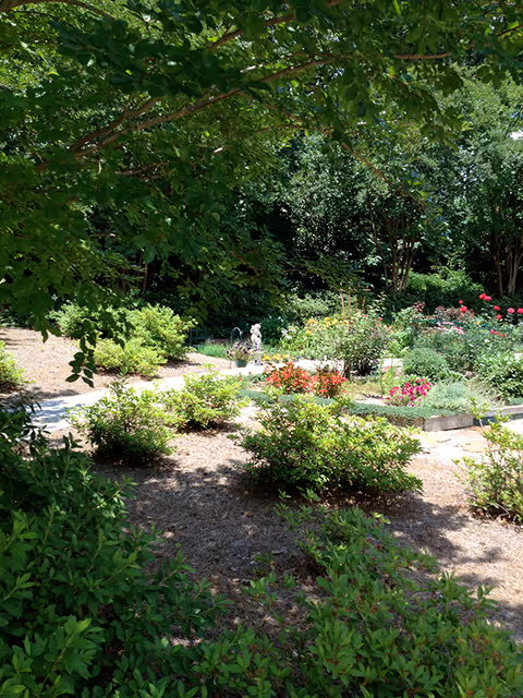 A lush garden area with various green shrubs, flowering plants, and trees providing shade. There is a small statue and a pathway winding through the garden beds filled with colorful flowers.