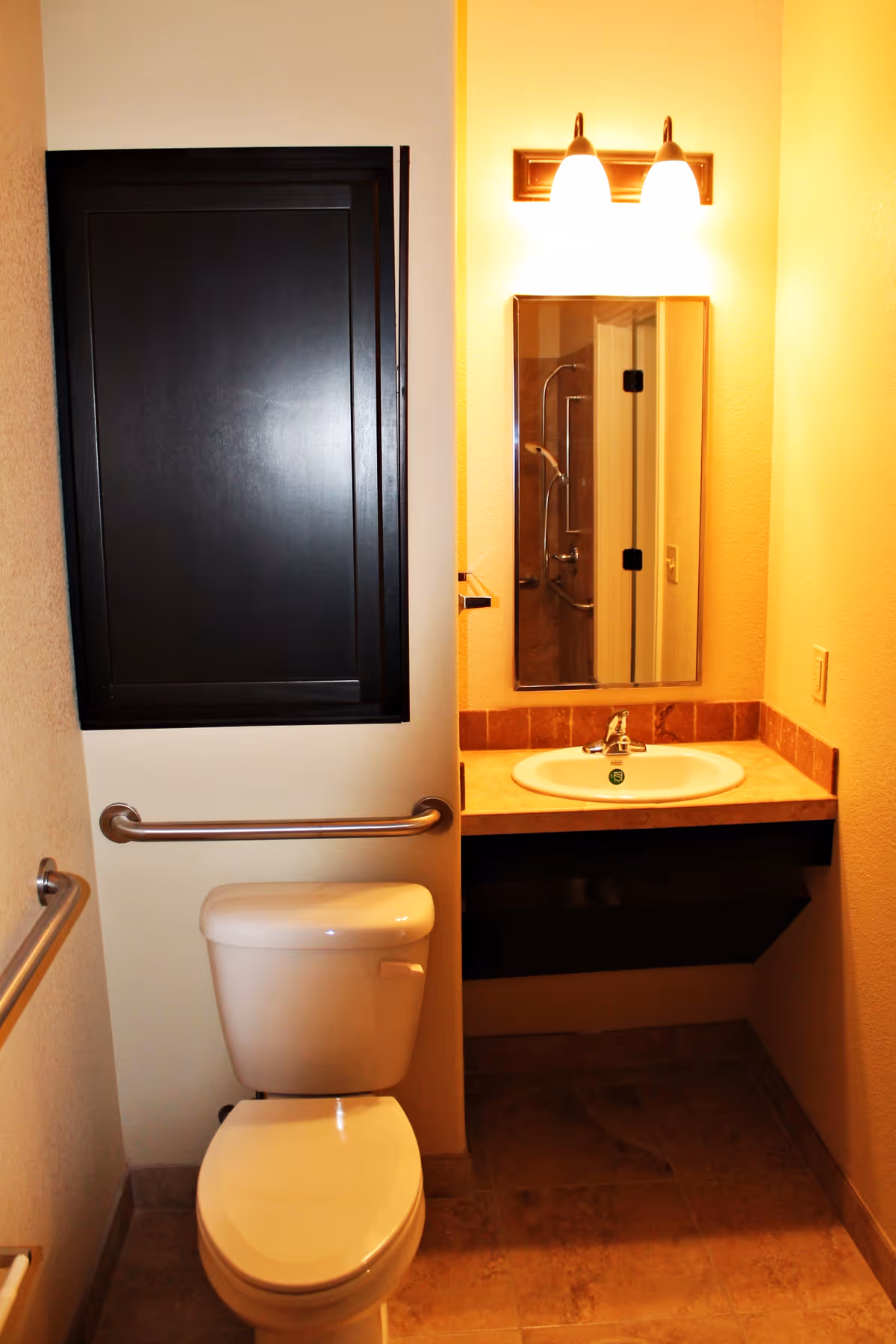 A small bathroom with a white toilet, a wall-mounted grab bar, a sink with a countertop, a mirror above the sink, and a light fixture with two bulbs above the mirror. There is a black cabinet mounted on the wall next to the toilet.