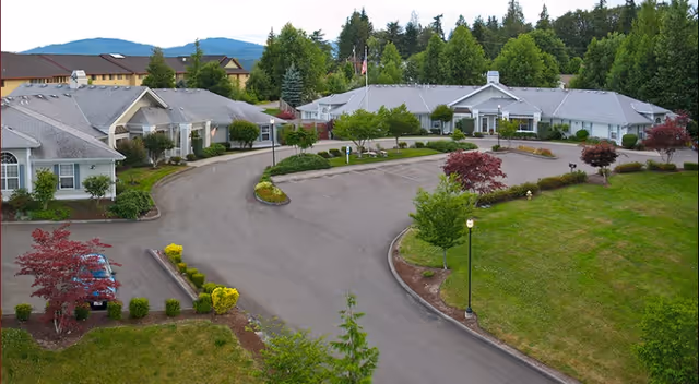 Aerial view of a senior living facility with multiple single-story buildings surrounded by landscaped greenery, trees, and a curved driveway. The area is well-maintained with parking spaces and a backdrop of forested hills under a clear sky.