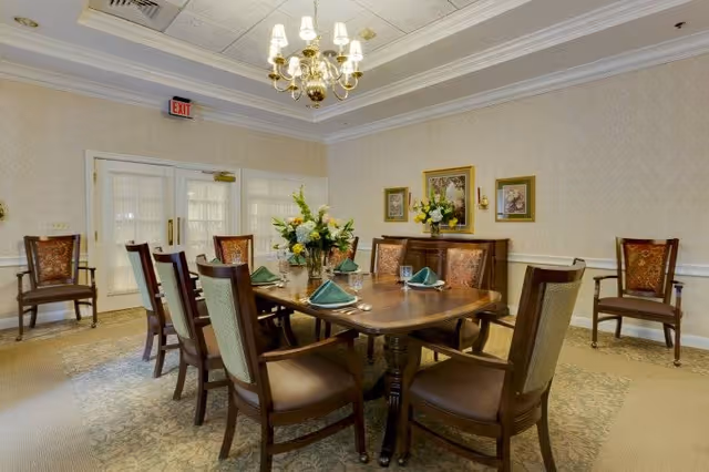 A formal dining room with a large wooden table set with green folded napkins, glasses, and silverware. The room features upholstered wooden chairs, floral centerpieces on the table and sideboard, framed artwork on the walls, a chandelier hanging from the ceiling, and double doors with glass panels.