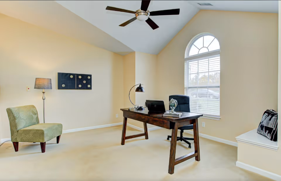 A bright office room with beige walls and carpet, featuring a wooden desk with a laptop, desk lamp, globe, and books. There is a black office chair behind the desk, a green upholstered chair to the left, a floor lamp, and a large arched window with white blinds letting in natural light. A ceiling fan with dark blades is mounted on the vaulted ceiling.
