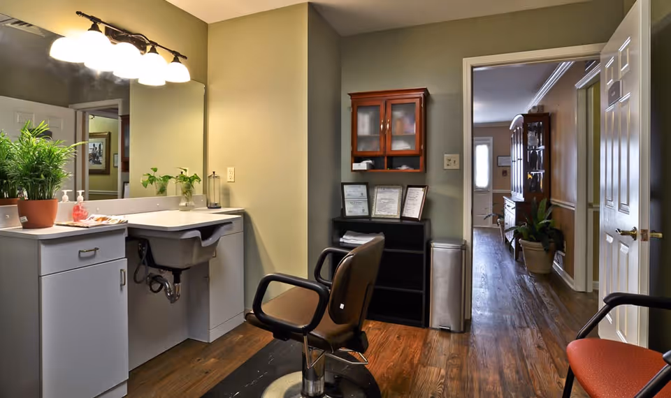 Interior view of a room with a sink and countertop on the left, a salon-style chair in the center, and a hallway leading to another room in the background. There are plants on the countertop and a cabinet with glass doors mounted on the wall. The floor is wooden, and the walls are painted in a light green color.