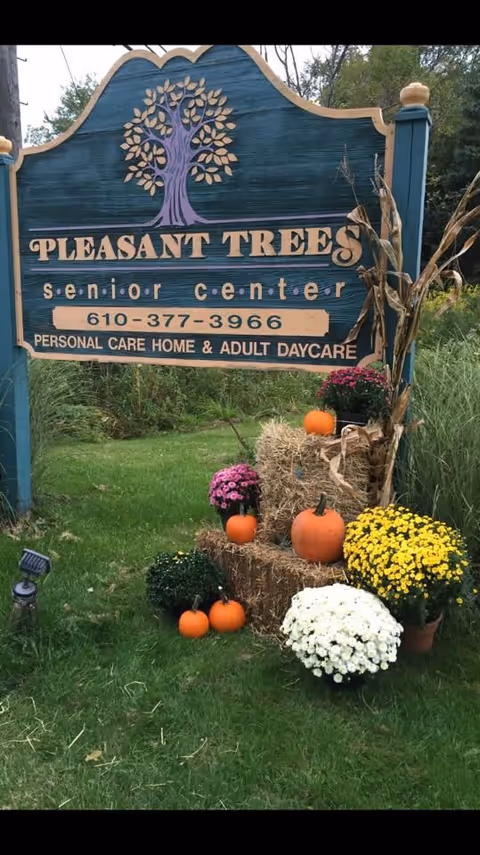 Outdoor wooden sign for Pleasant Trees Senior Center, a personal care home and adult daycare, surrounded by hay bales, pumpkins, and colorful potted flowers on a grassy area.