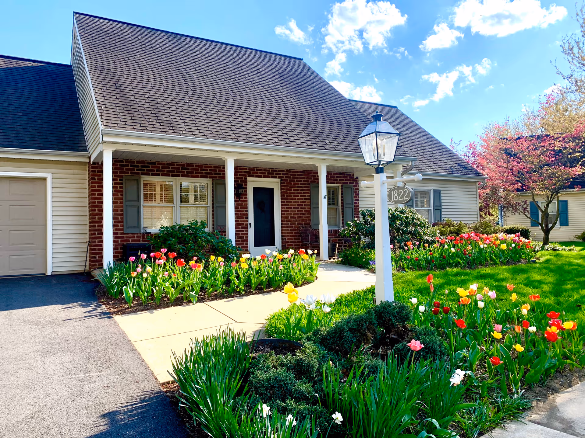 A single-story residential building with a brick and siding exterior, a dark shingled roof, and a white front door. The house has a small porch supported by white columns and is surrounded by a well-maintained garden with colorful tulips and green shrubs. A white lamp post with the number 1822 is visible in the front yard. The sky is blue with scattered clouds.