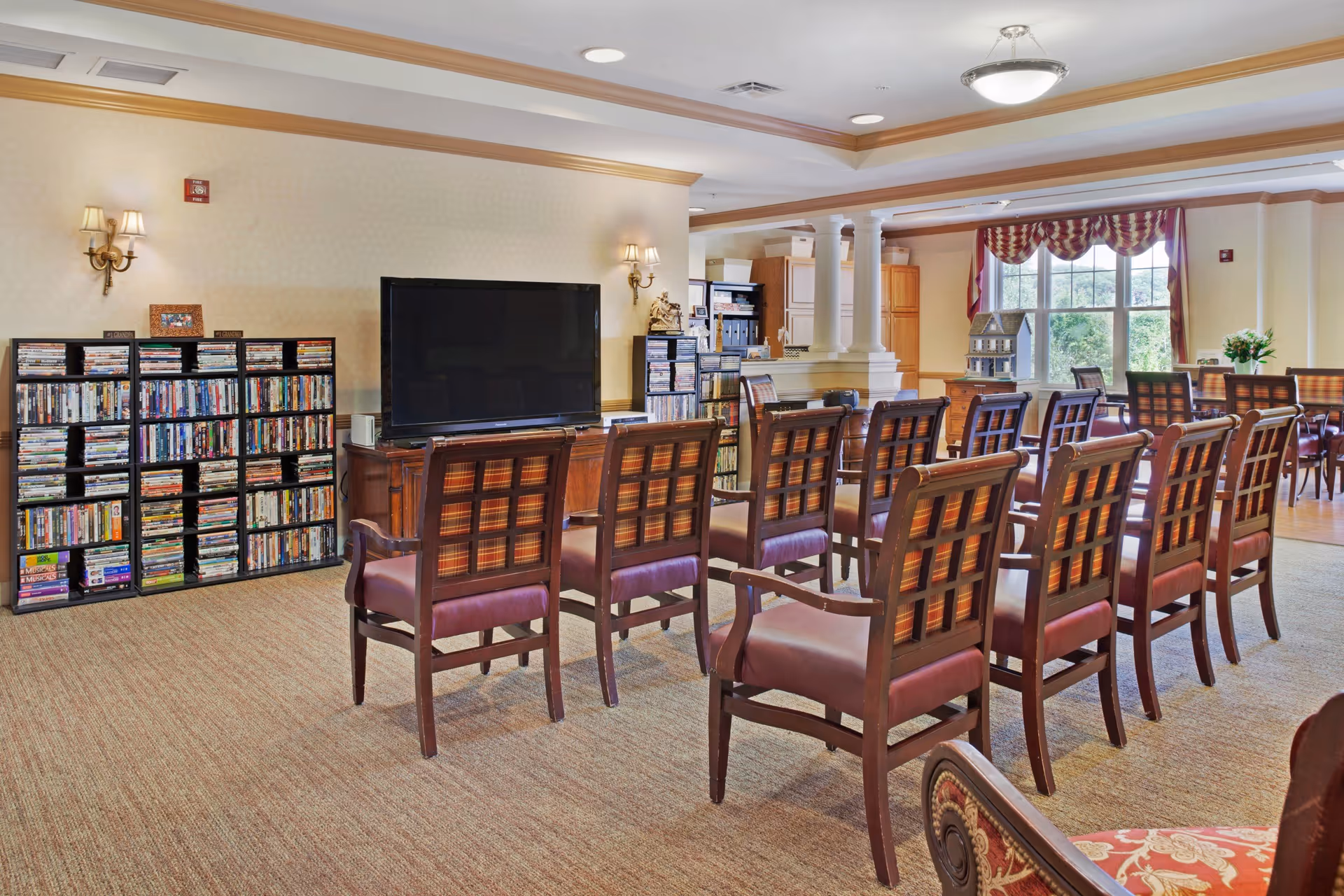 A communal activity room with rows of wooden chairs facing a large TV, shelves of DVDs, and windows letting in natural light.