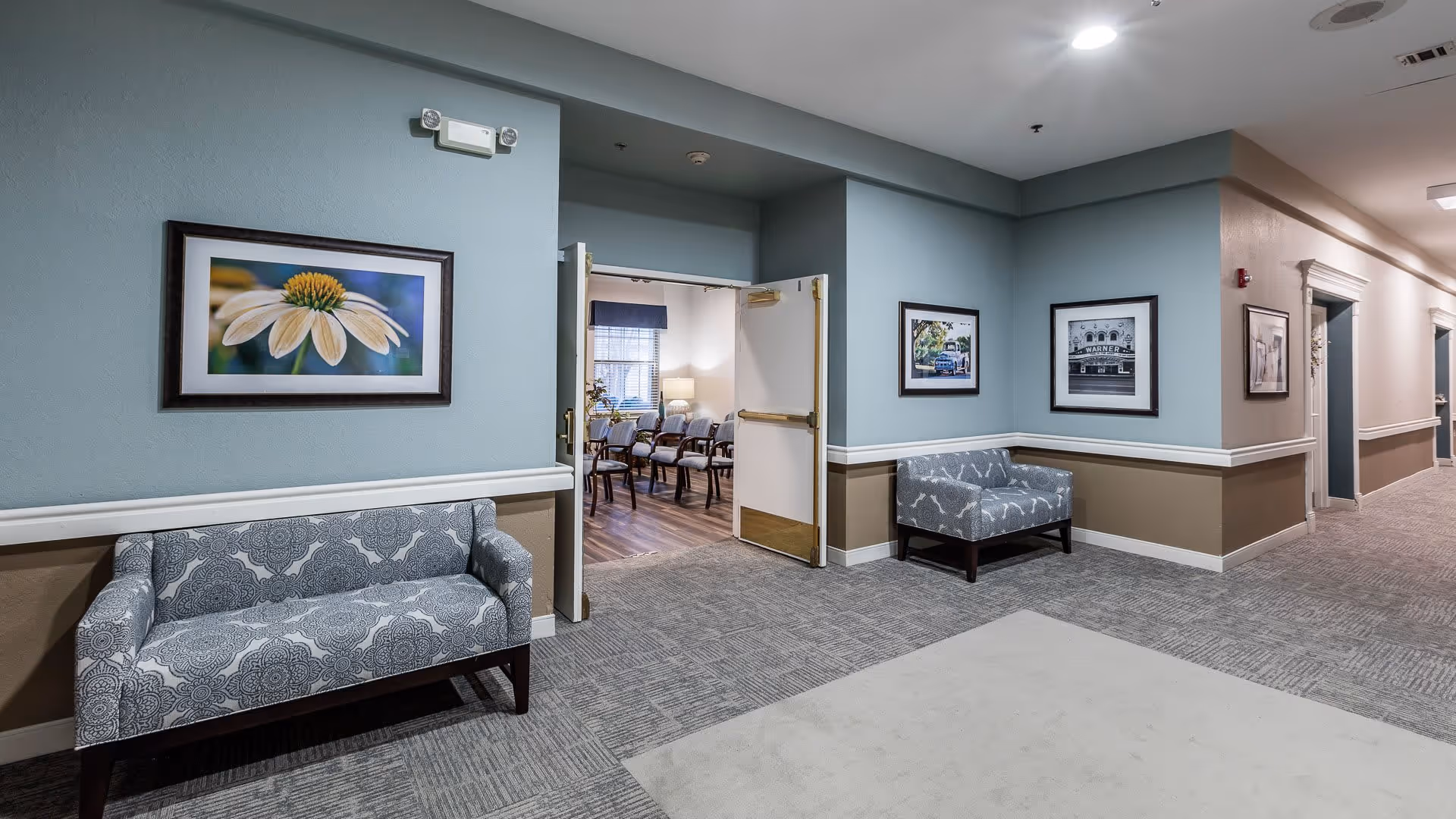 A hallway in an assisted living facility with patterned armchairs against the walls, framed pictures hanging above the chairs, and an open door leading to a room with multiple chairs and a window with blinds. The walls are painted in two tones with a chair rail, and the floor is carpeted with a light pattern.