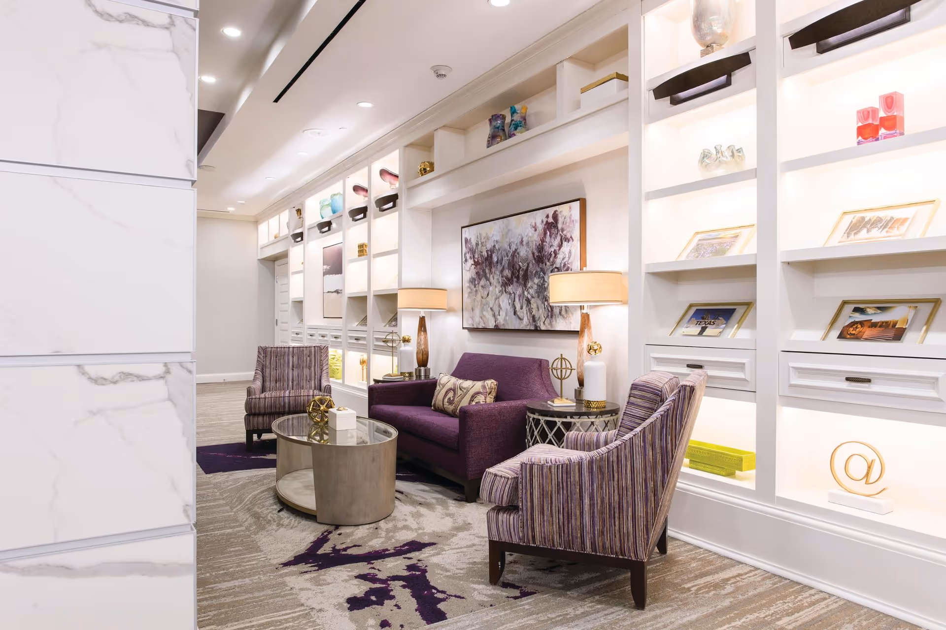 A modern and elegant lounge area with a purple sofa, two striped armchairs, a round glass coffee table, and two side tables with lamps. The back wall features built-in white shelves with decorative items and framed pictures, illuminated by soft lighting. The floor has a patterned carpet with purple accents.