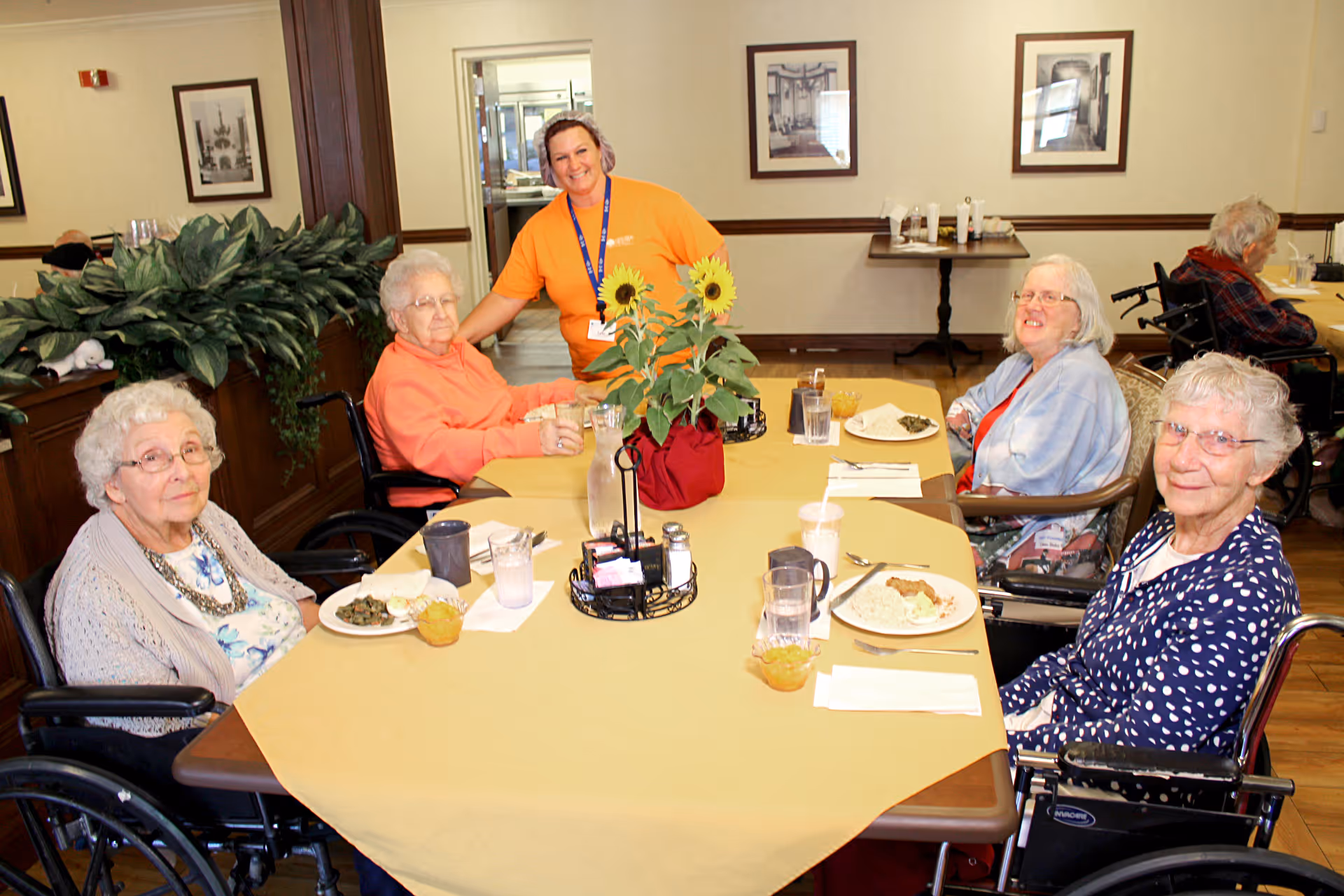 Five elderly women seated at a dining table with plates of food while a smiling staff member stands behind them.