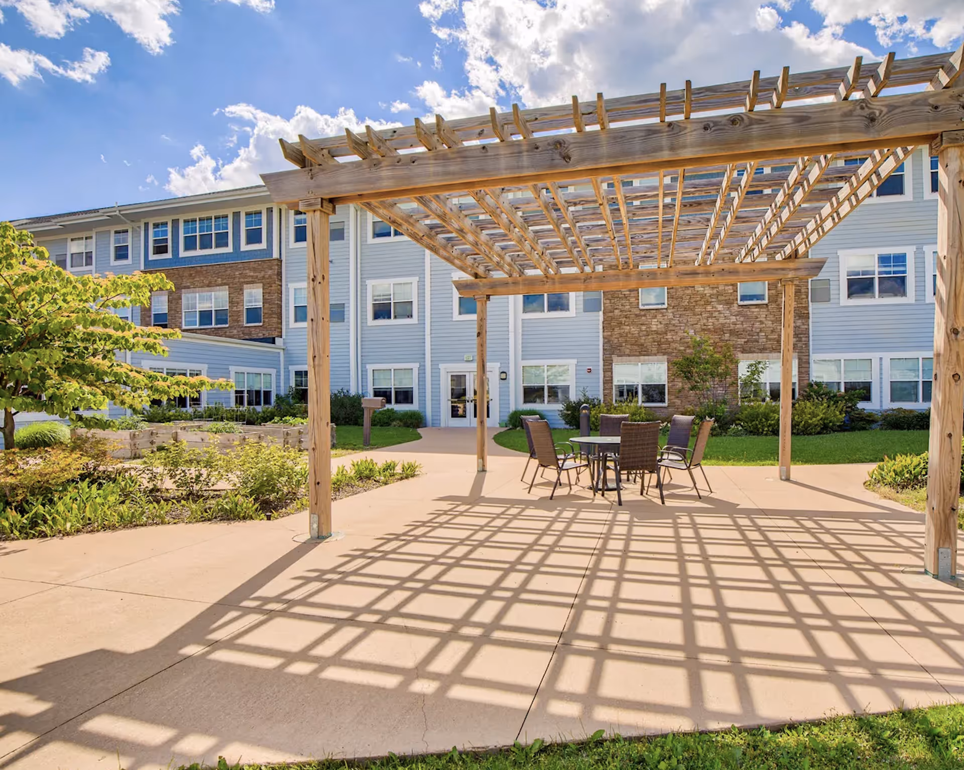 Outdoor patio area at StoryPoint Schererville featuring a wooden pergola casting a grid-like shadow on the concrete ground. Under the pergola, there is a round table with six chairs. The background shows a multi-story building with light blue siding and stone accents, surrounded by greenery and a clear blue sky with some clouds.