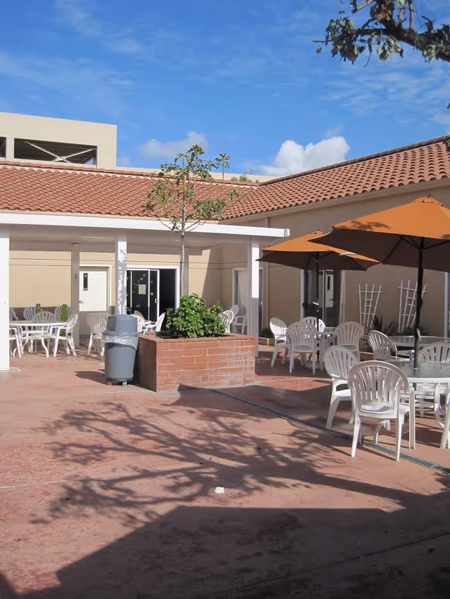 Outdoor courtyard area with several white plastic chairs and tables, some shaded by large orange umbrellas. There is a small tree planted in a brick planter in the center, and the building surrounding the courtyard has beige walls and a red tile roof. The sky is clear and blue with a few clouds.