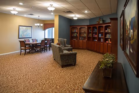 A spacious senior living common area featuring a patterned carpet, a wooden dining table with six chairs, two upholstered armchairs, a large wooden bookshelf filled with books, and framed artwork on the walls. The room is well-lit with ceiling lights and natural light from a window.