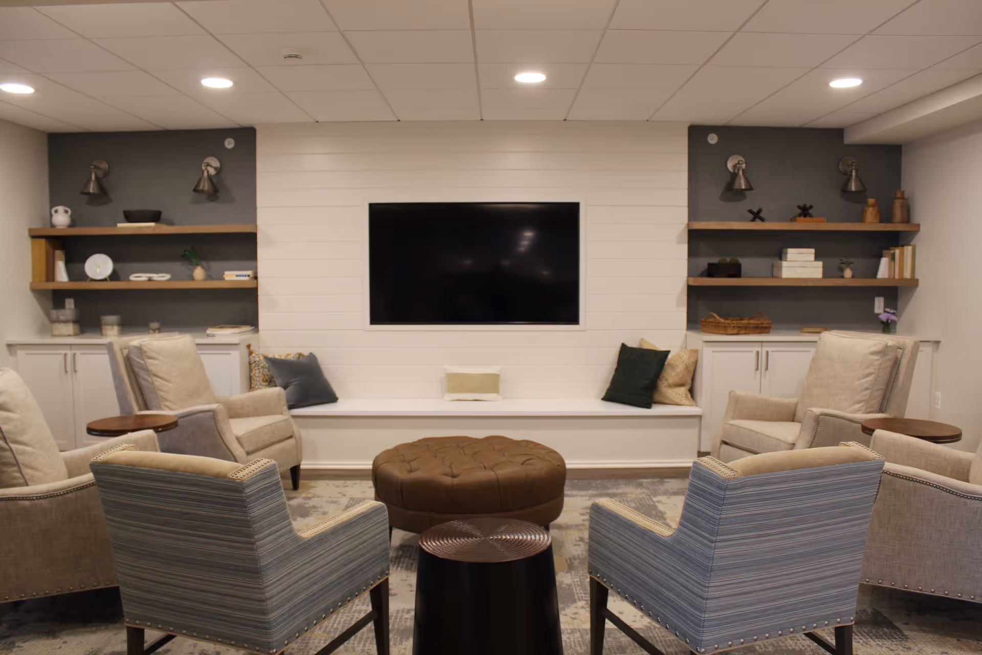 A cozy living room with a large flat-screen TV mounted on a white shiplap wall. The room features six upholstered chairs arranged in a circle around a round brown ottoman and a small round side table. On either side of the TV, there are built-in shelves with decorative items and cabinets below. The lighting is recessed in the ceiling, creating a warm and inviting atmosphere.