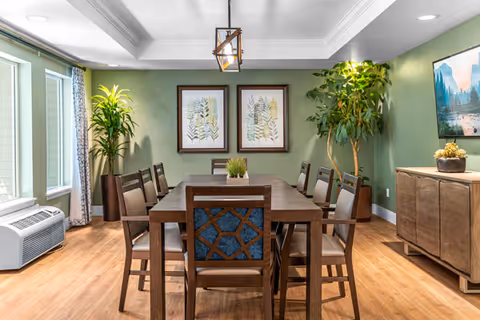 A well-lit dining room with a wooden table surrounded by eight chairs. The walls are painted green and decorated with two framed botanical prints. There are two large potted plants in the corners, a wooden sideboard with a decorative item, and a modern light fixture hanging from the ceiling. Large windows with curtains allow natural light to fill the room.