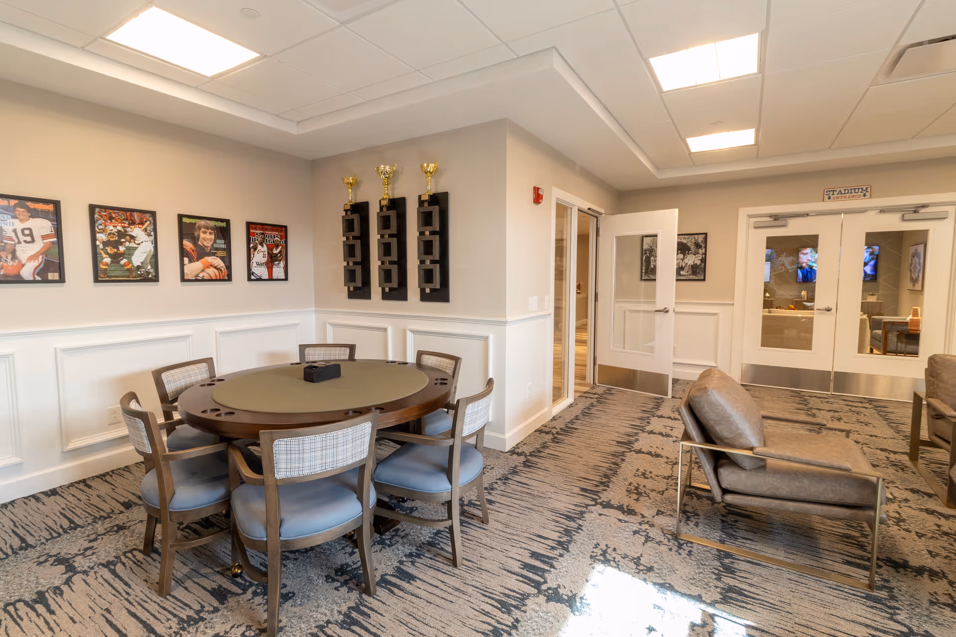 A well-lit common area with a round table surrounded by six chairs in the foreground. The walls are decorated with framed sports photos and three trophy displays. To the right, there are two leather armchairs and double glass doors leading to another room with a television visible inside.