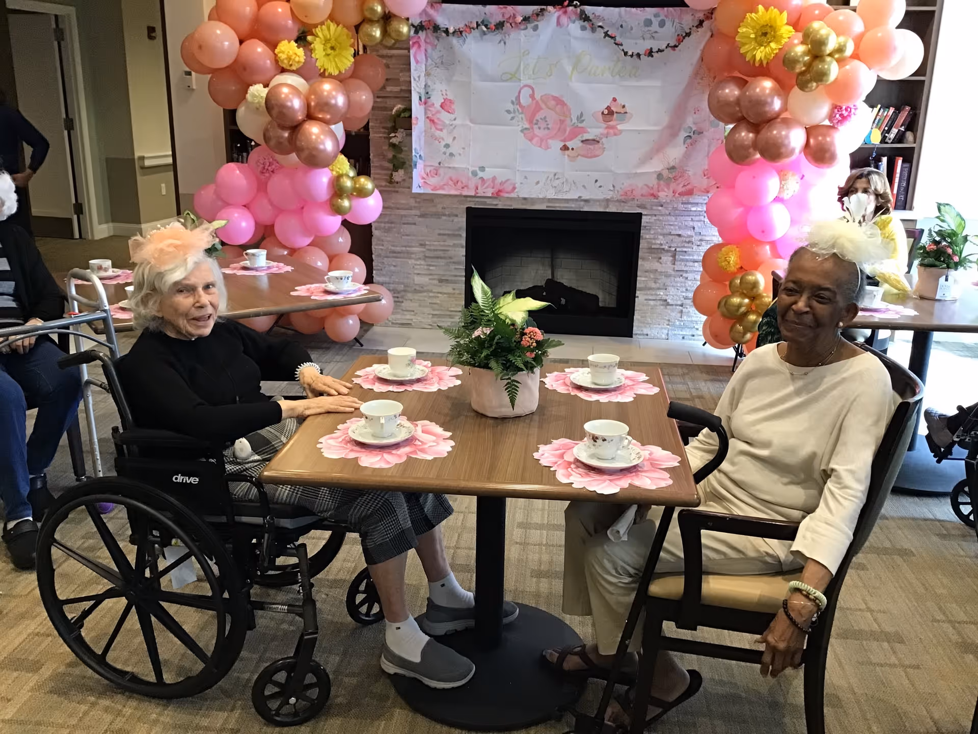 Two elderly women sitting at a table in a decorated room, each wearing a festive hat. The table is set with teacups and floral placemats, and there is a flower arrangement in the center. Behind them is a fireplace with a banner and balloon decorations in shades of pink, gold, and peach. Other people are visible in the background seated at tables.
