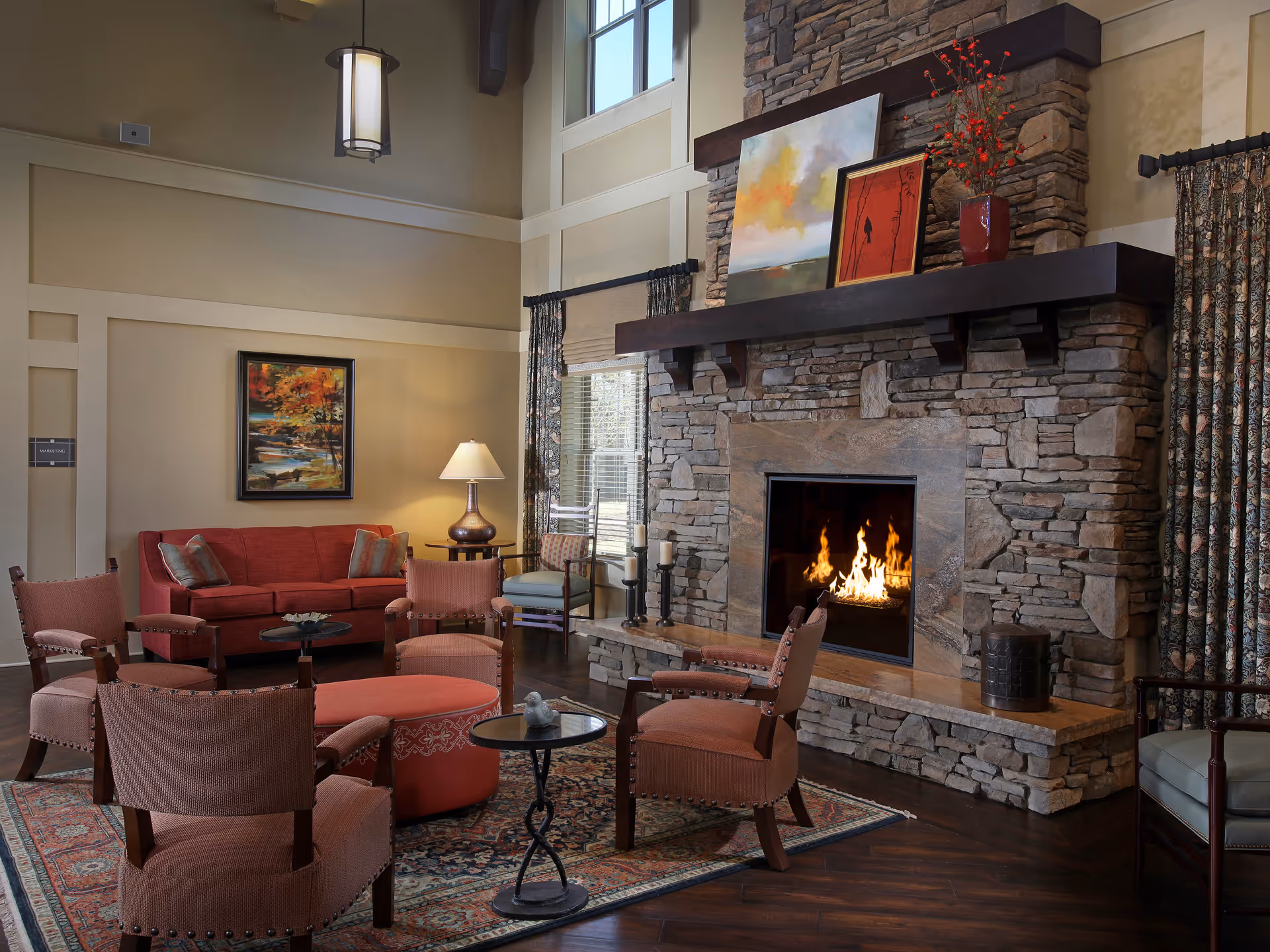 A cozy living room with a stone fireplace featuring a burning fire. The room has a red sofa, four upholstered chairs arranged around a round ottoman, a side table with a lamp, and a large window with patterned curtains. There are paintings and a vase with red flowers on the fireplace mantel.