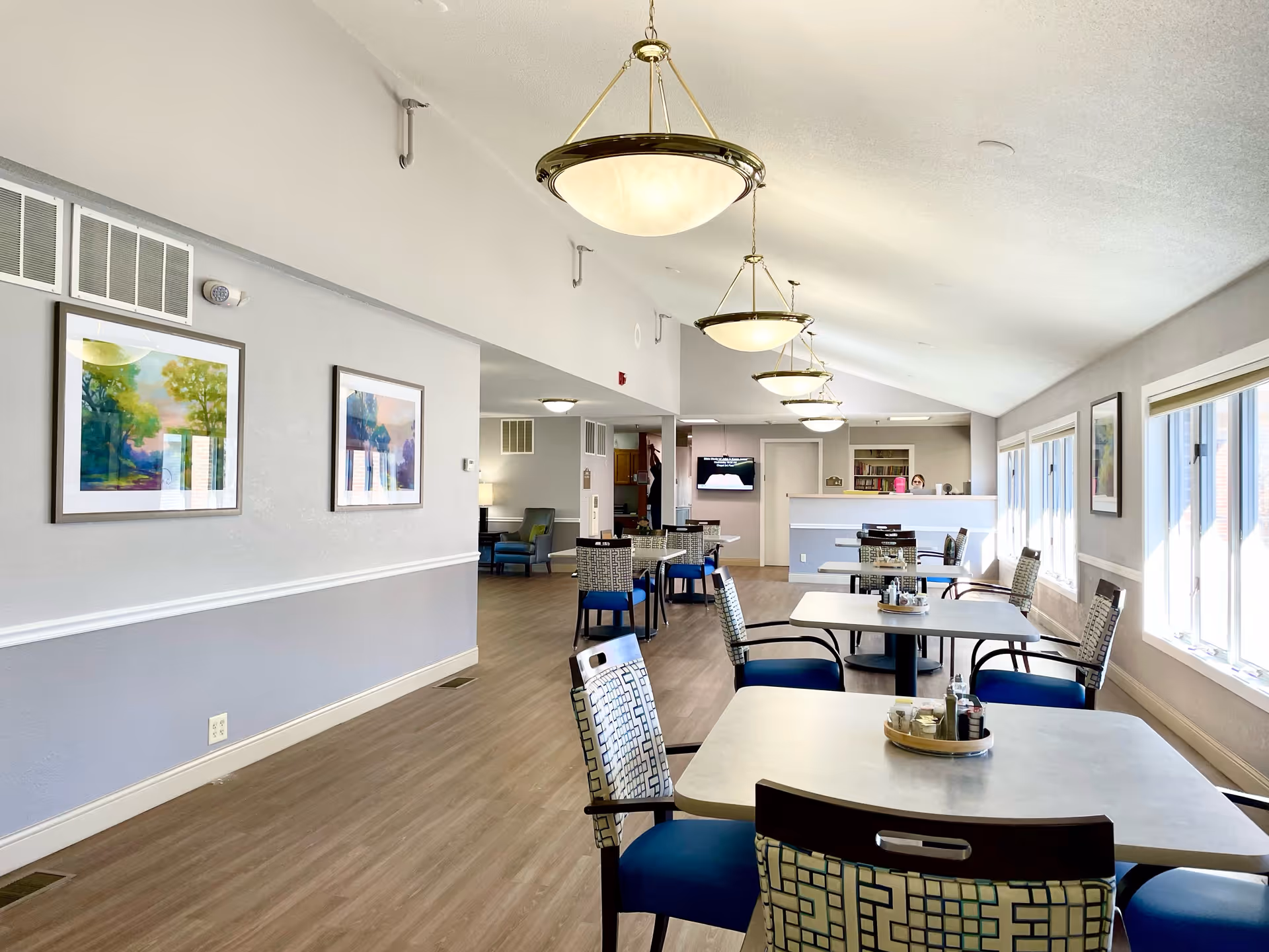 Bright communal dining area with several tables and patterned chairs, pendant lights, and large windows in a senior living facility.