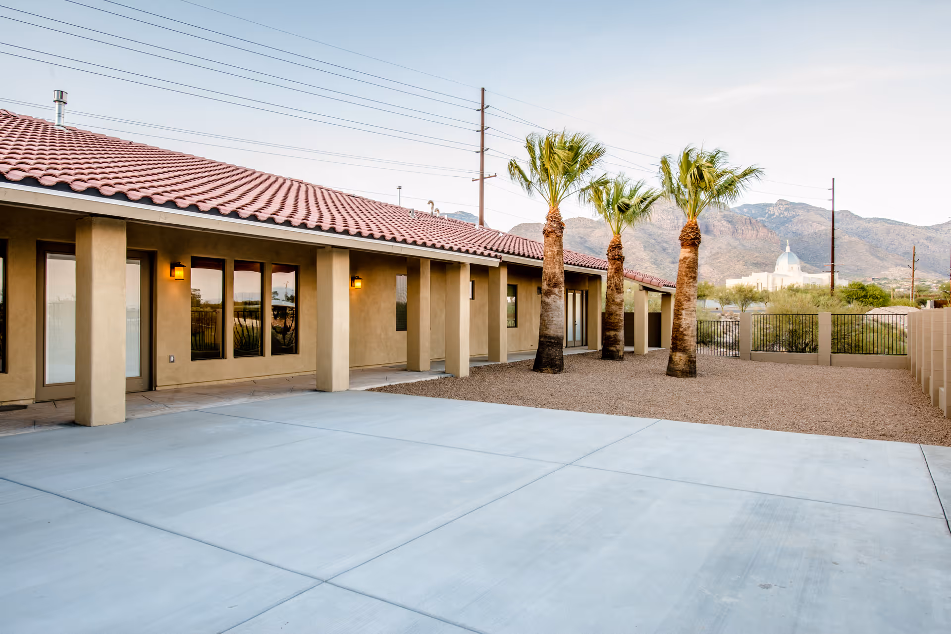 Single-story stucco building with a red tile roof, palm trees, and a paved courtyard set against distant mountains.
