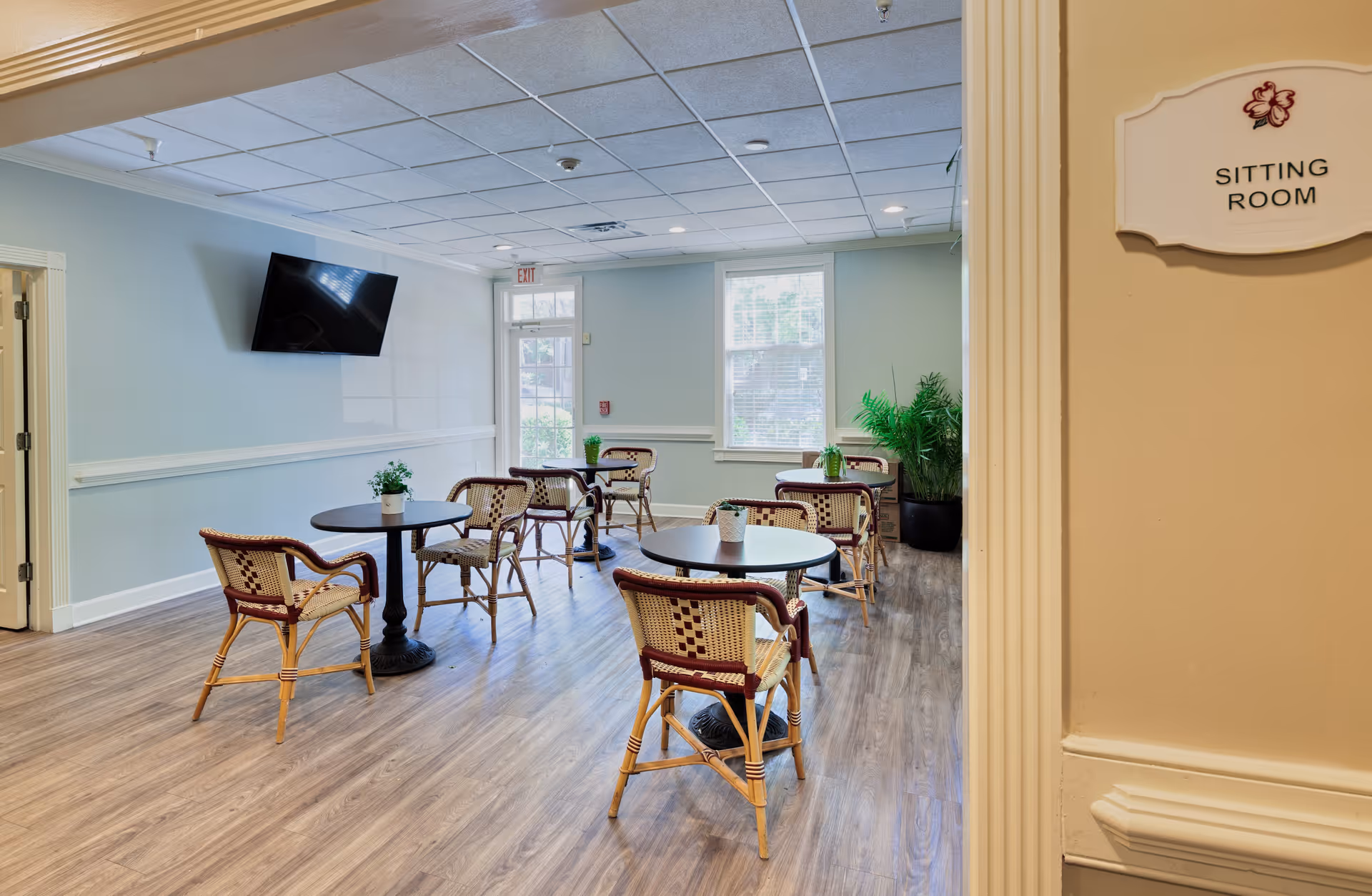 A sitting room with several round tables and wicker chairs arranged on a wood floor. The walls are painted light blue with white trim, and there is a flat-screen TV mounted on one wall. A window and a glass door allow natural light into the room. A sign on the wall near the entrance reads 'SITTING ROOM'.