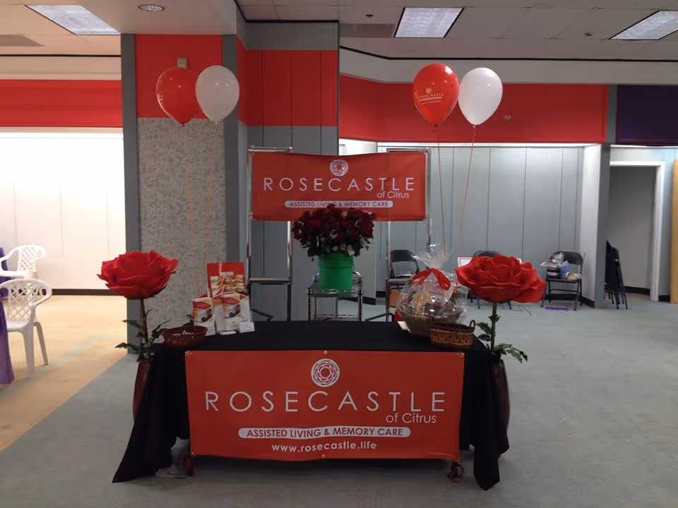 A promotional table setup for Rosecastle of Citrus assisted living and memory care, featuring a black tablecloth with a red banner, two large artificial red roses in vases on each side, red and white balloons, brochures, and a gift basket. The background shows a room with chairs and a red and gray wall.