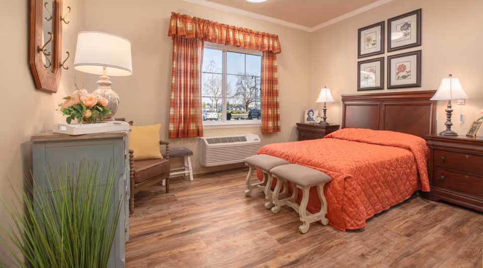 A cozy bedroom with a wooden bed frame and an orange quilted bedspread. Two cushioned benches are placed at the foot of the bed. There are two wooden nightstands on either side of the bed, each with a lamp and framed photos. Above the bed are four framed floral prints. A window with red and orange plaid curtains lets in natural light. A wooden chair with a yellow cushion and a small gray stool are near a light blue dresser with a lamp and flower arrangement on top. The room has wood flooring and beige walls.
