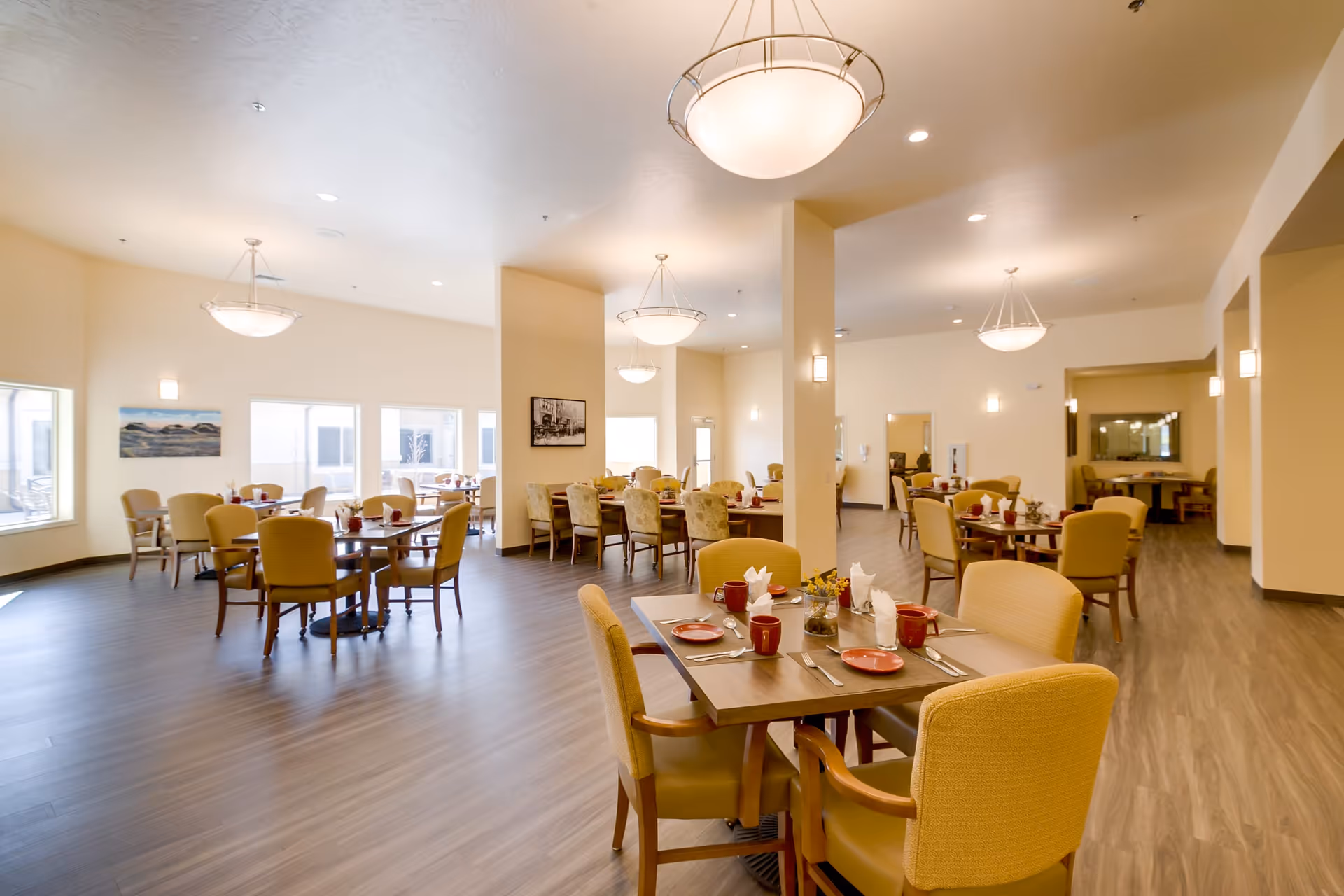 A spacious senior living dining room with multiple tables set with red plates, cups, silverware, and napkins. The room features beige walls, large windows letting in natural light, wooden flooring, and modern ceiling lights. Comfortable upholstered chairs surround each table.