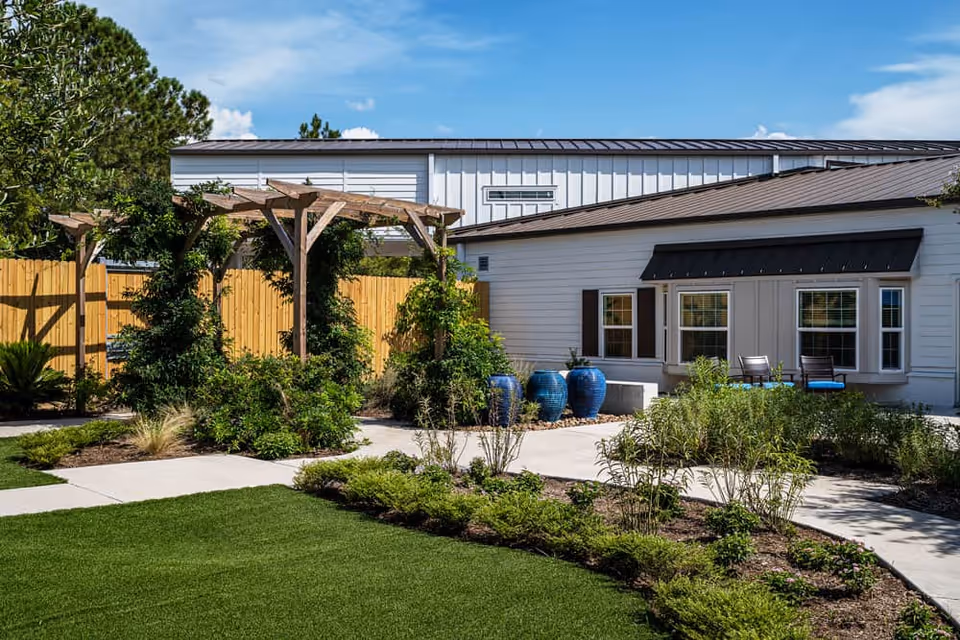 Outdoor garden area at Reunion Court of Clear Lake featuring a wooden pergola with climbing plants, a well-maintained lawn, landscaped bushes and shrubs, a concrete walkway, and a white building with windows and two chairs on a small patio.