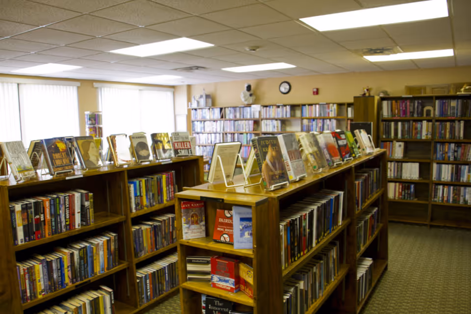 Interior view of a library room with wooden bookshelves filled with books. Some books are displayed facing forward on top of the shelves. The room has large windows with vertical blinds allowing natural light to enter. A clock and a bust are visible on the back wall above more bookshelves.