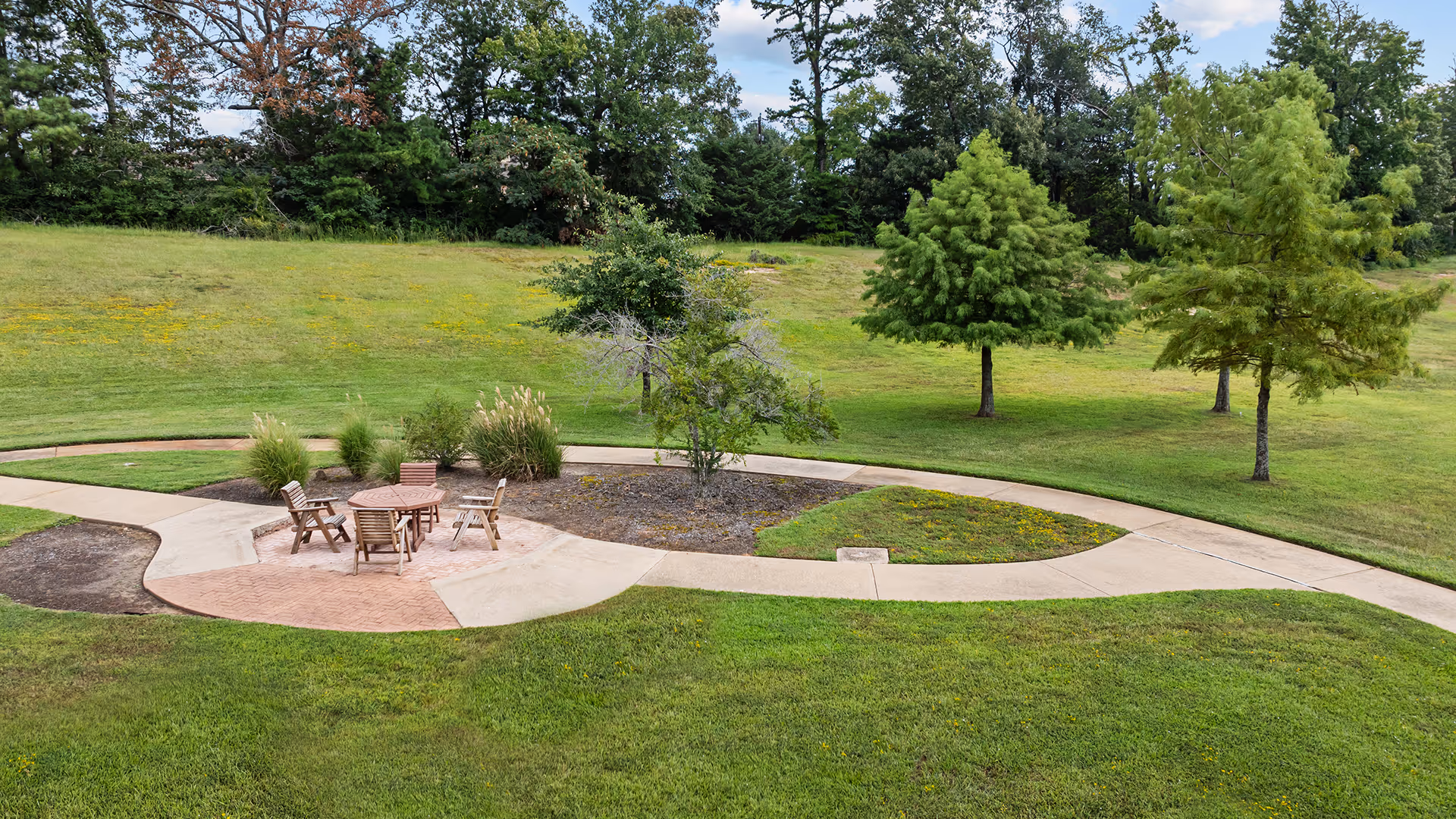 Outdoor seating area with a round wooden table and four wooden chairs on a paved circular patio surrounded by grass, trees, and a curved concrete walkway in a park-like setting.