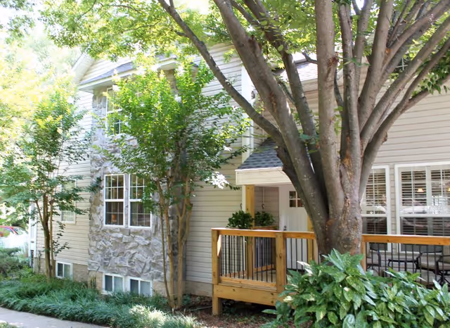 Exterior view of a residential-style building with beige siding and stone accents, surrounded by trees and greenery. There is a wooden porch with railing and a large tree in the foreground.