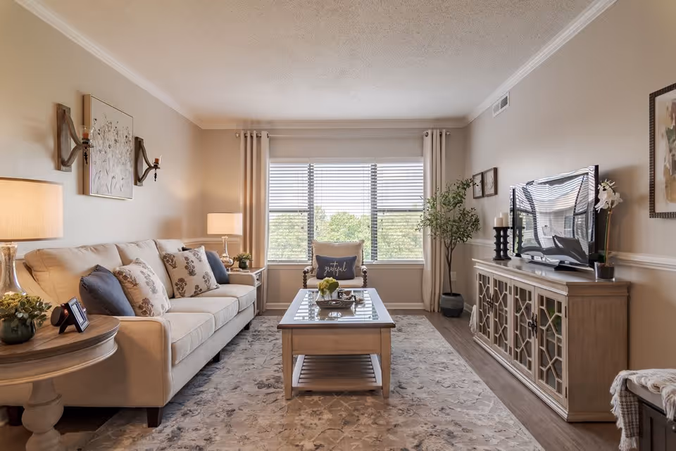 A cozy living room with a beige sofa adorned with floral and solid pillows, a wooden coffee table with decorative items, a large window with white curtains letting in natural light, a wooden TV stand with a flat-screen television, and a potted plant in the corner. The room has neutral-colored walls and a patterned area rug covering the wooden floor.