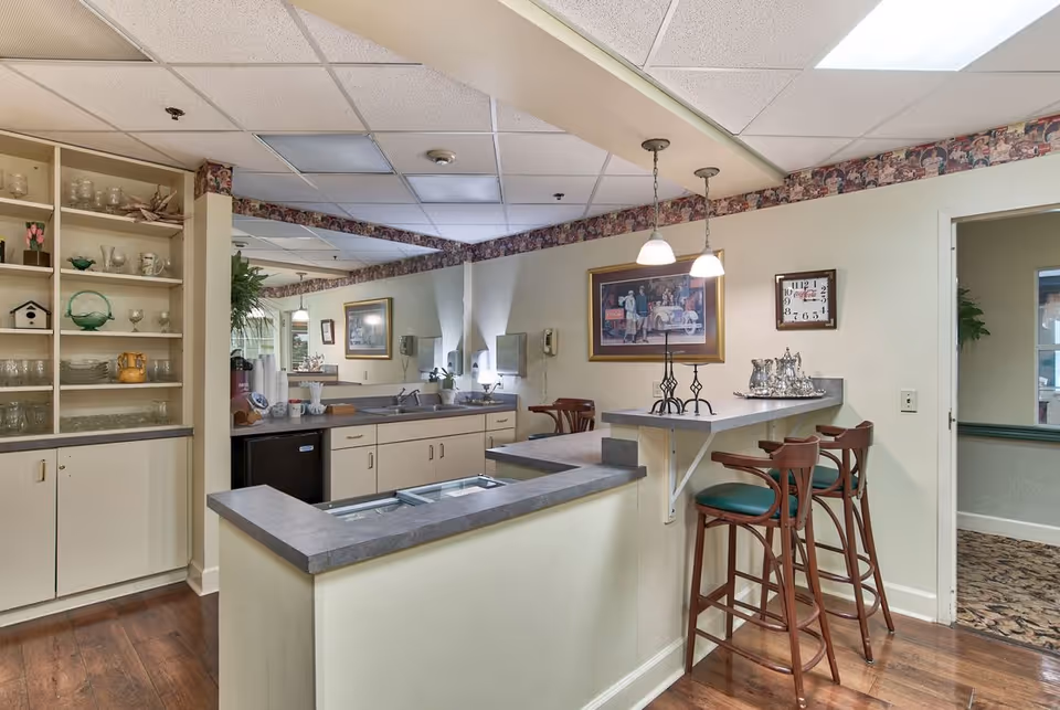 Interior view of a kitchen area in an assisted living facility featuring a countertop with two high chairs, a sink, cabinets, shelves with glassware and decorative items, pendant lights, a wall clock, and framed artwork on the walls.