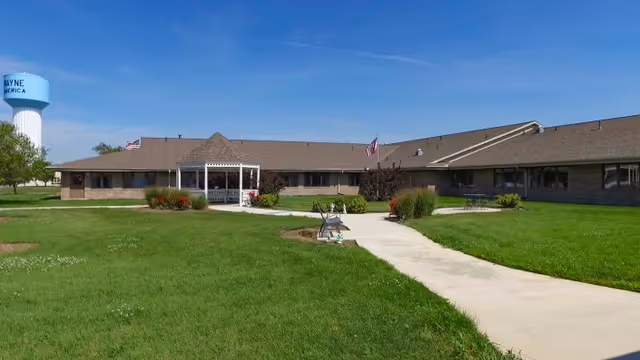 Single-story brick care facility with a covered entrance, concrete walkway and green lawn under a blue sky.