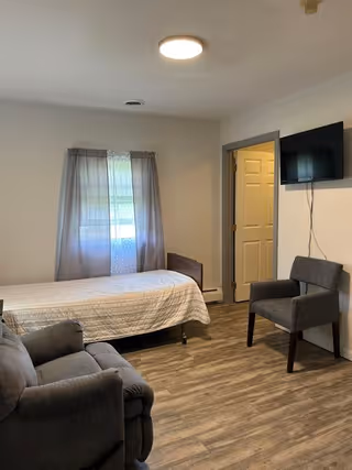 Furnished bedroom with a single bed, two chairs, a wall-mounted TV, and a window with curtains.