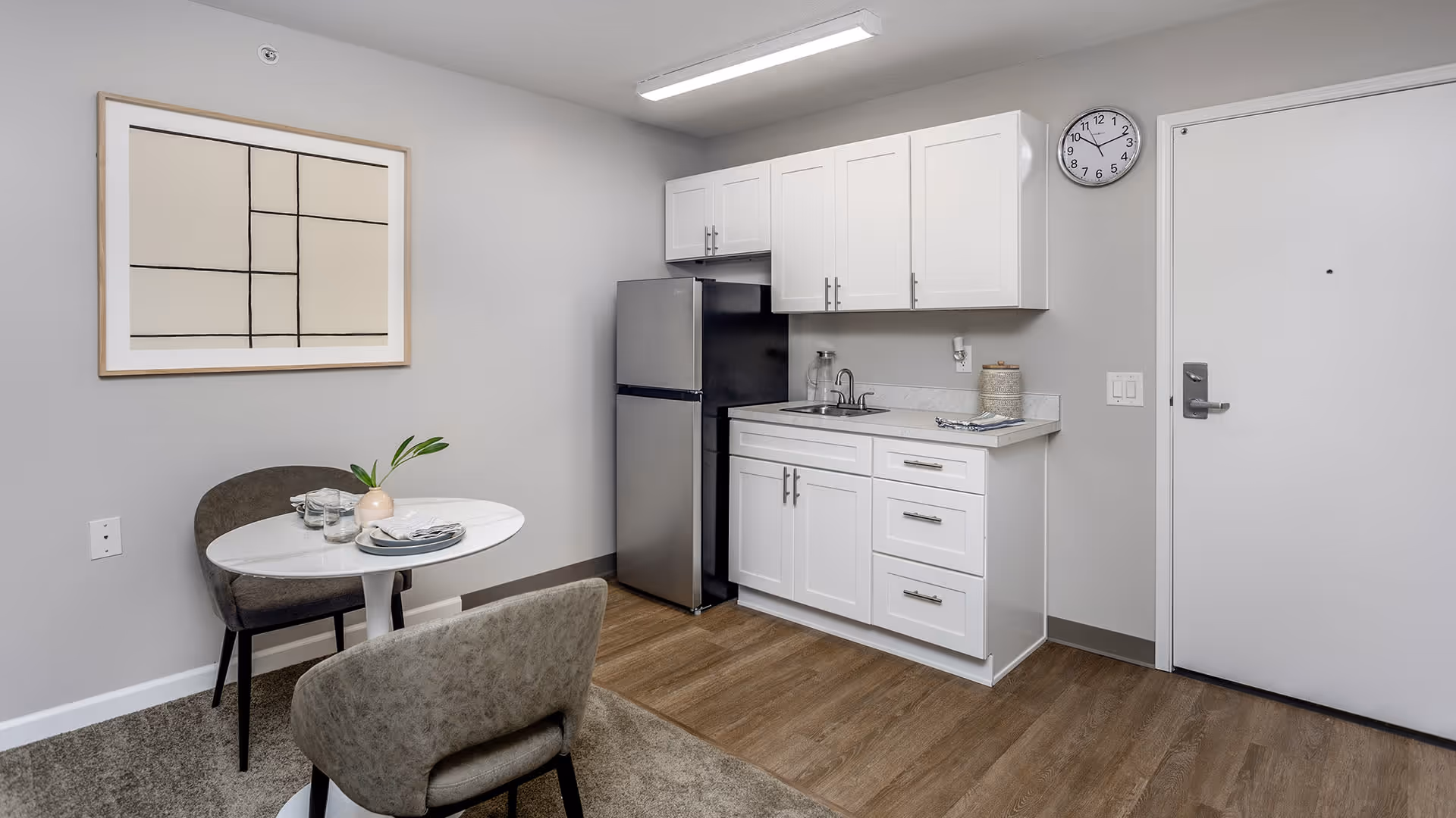 A small kitchenette area with white cabinets, a stainless steel refrigerator, a sink, and a countertop. Next to the kitchenette is a small round dining table set for two with gray upholstered chairs. A modern abstract painting hangs on the wall above the table. The floor is a combination of wood and carpet, and a wall clock is mounted near a closed white door.
