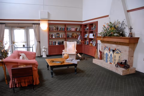 A cozy living room with a pink sofa, a patterned armchair, a wooden coffee table, and a fireplace decorated with miniature house models and a toy car. There are built-in red bookshelves filled with books and decorative items, a lamp, and a statue near a glass door with curtains.