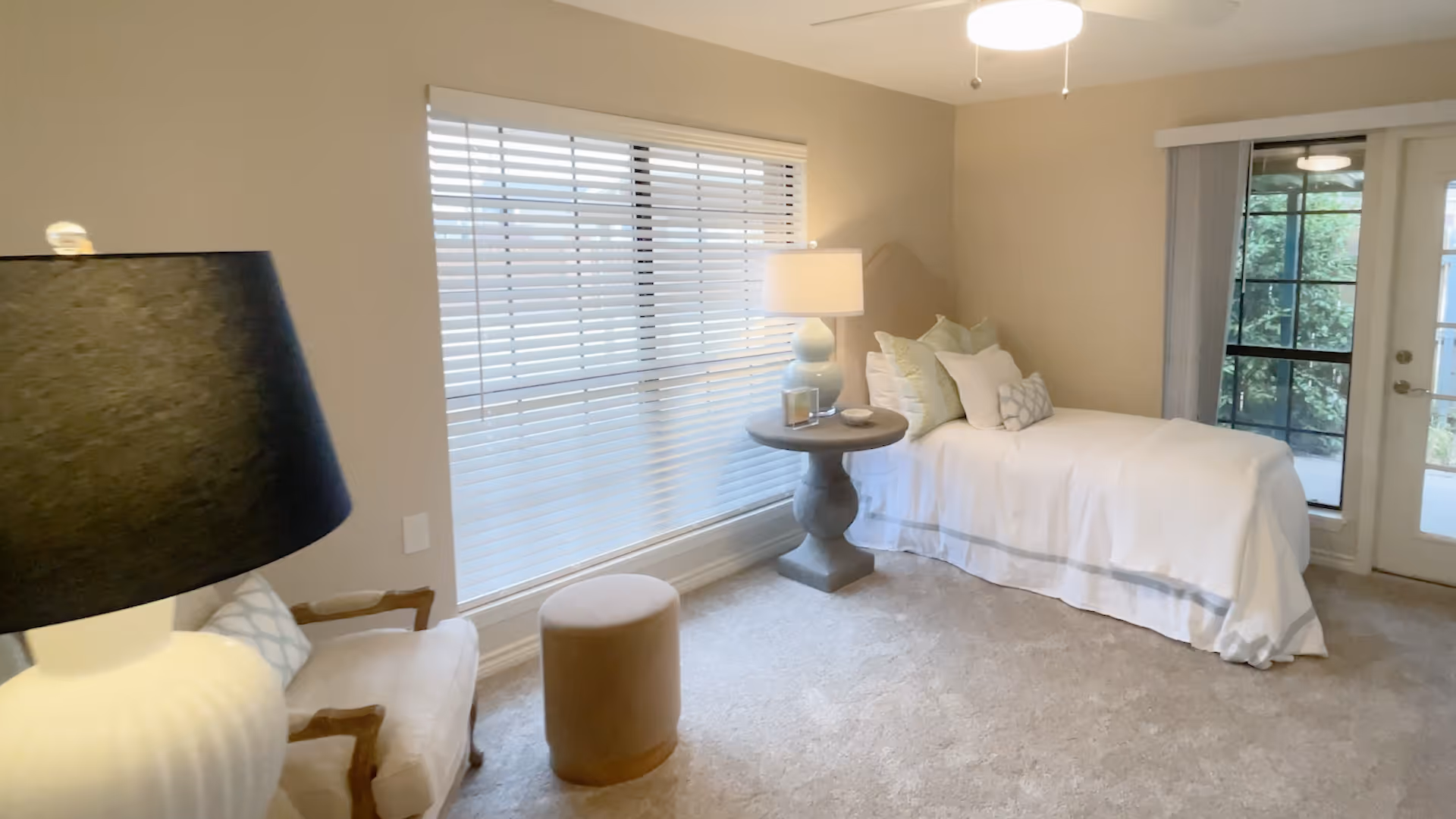 Well-lit bedroom with a single bed, round bedside table and lamp, an armchair, ottoman, and large window with blinds.