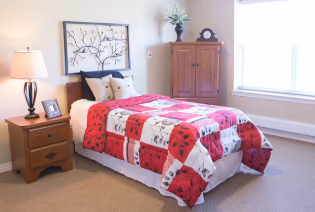 A cozy bedroom with a single bed covered by a red, white, and black patterned quilt. The bed has several pillows and a decorative metal wall art piece above the headboard. To the left of the bed is a wooden nightstand with a lamp and a framed photo. To the right is a wooden cabinet with a clock and a vase with flowers on top. A large window with blinds allows natural light into the room.