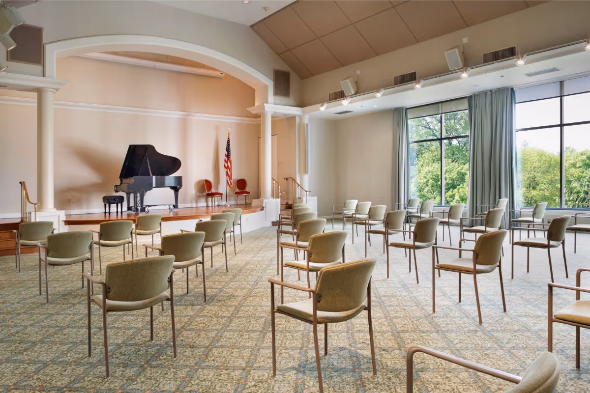 A spacious room with rows of green cushioned chairs arranged facing a small stage. On the stage, there is a black grand piano, two red chairs, and an American flag. Large windows on the right side let in natural light and show green trees outside. The room has a high ceiling with spotlights and speakers mounted.
