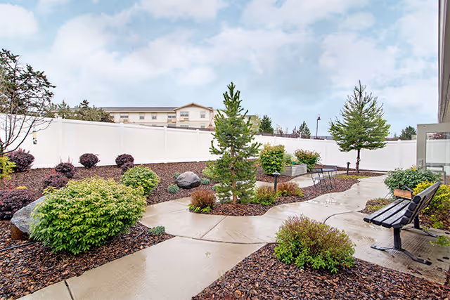 A landscaped outdoor garden area with a concrete pathway winding through various green shrubs, small trees, and mulch beds. There is a black bench on the right side and a white fence enclosing the area. A building is visible in the background under a partly cloudy sky.