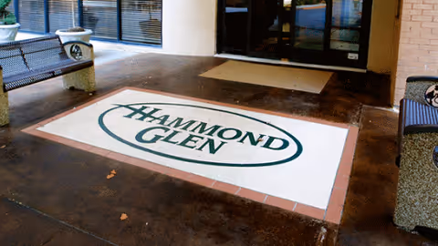 Entrance area of Hammond Glen Retirement Community featuring a large floor mat with the facility's name and logo. There are benches on either side and glass doors leading inside the building.