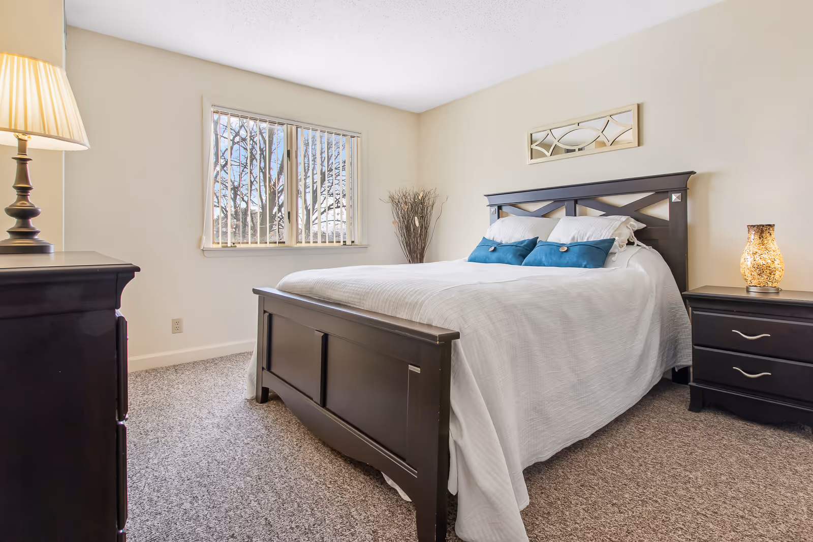 A bright bedroom with a large dark wooden bed frame, white bedding, and two blue accent pillows. There is a window with vertical blinds letting in natural light, a dark wooden dresser with a lamp on the left, and a matching nightstand with a decorative lamp on the right. The walls are light-colored and there is a decorative mirror above the bed.