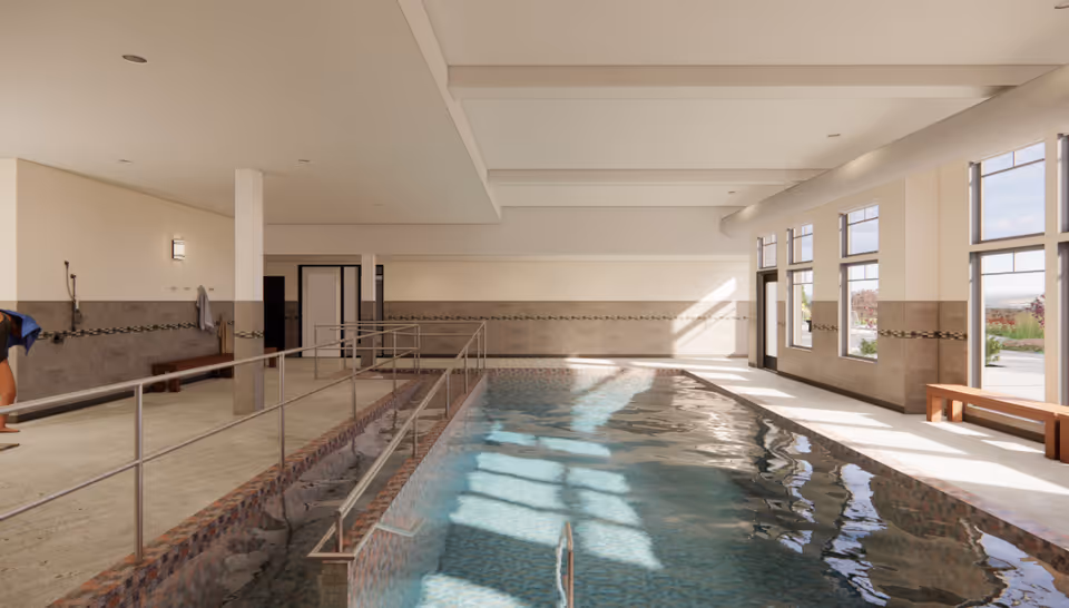 Indoor swimming pool area with handrails for accessibility, large windows letting in natural light, tiled walls, benches along the side, and a person bending down near the pool edge.