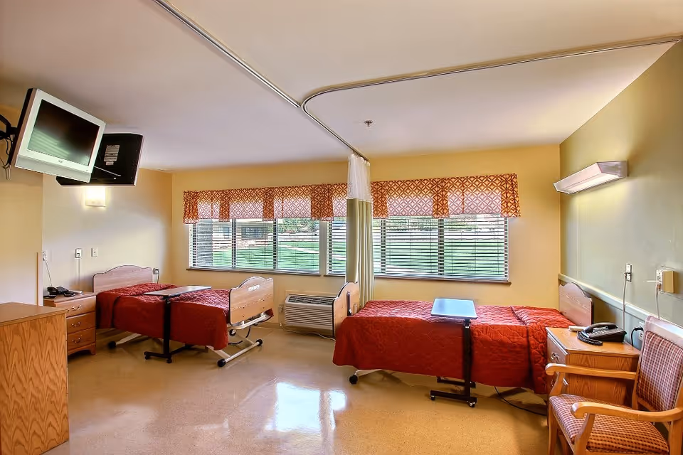 Two-bed patient room in a senior living facility with red bedspreads, a wall-mounted TV, bedside tables, chairs and a privacy curtain in front of large windows.